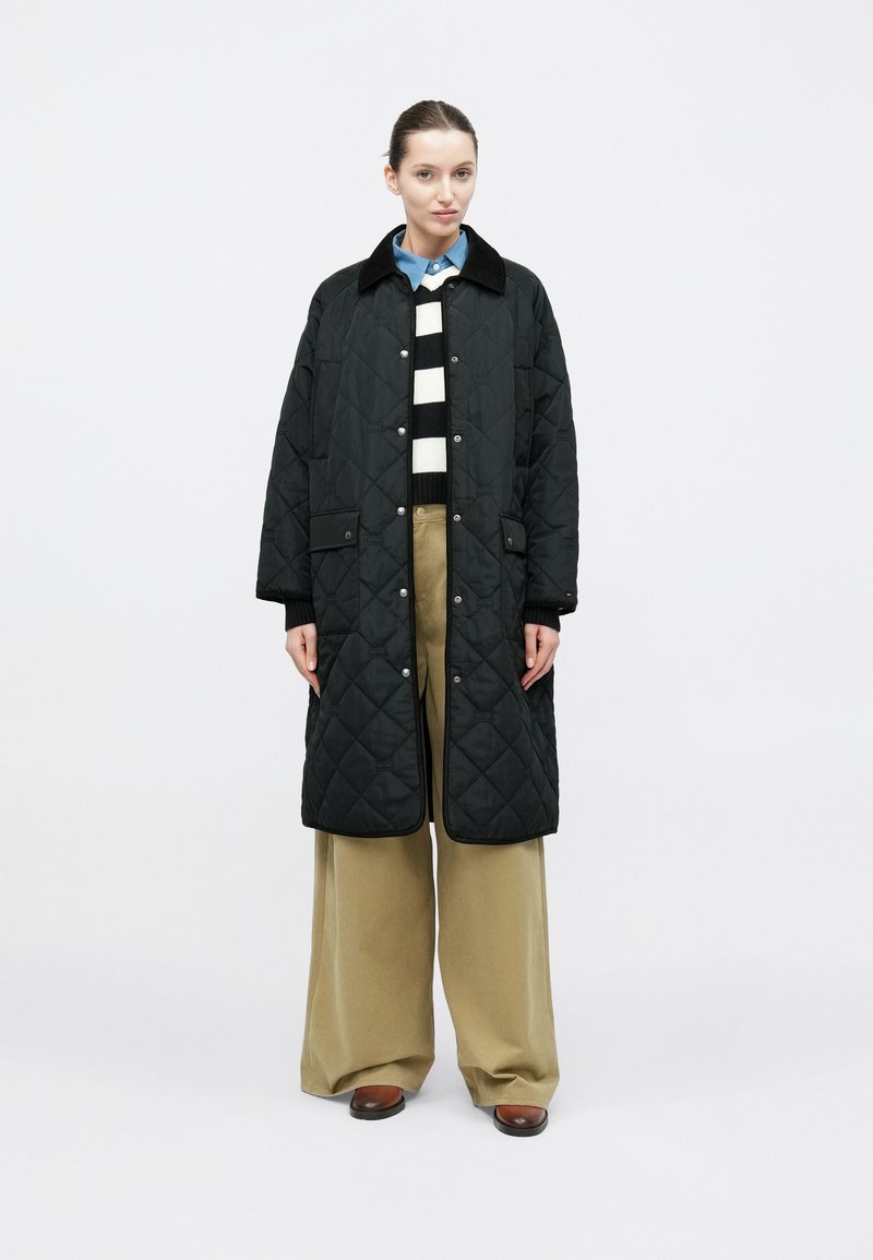 Čierny prešívaný dlhý kabát s golierom, nosený cez pruhovaný sveter a široké opálene hnedé nohavice, s patentkami a bočnými vreckami.