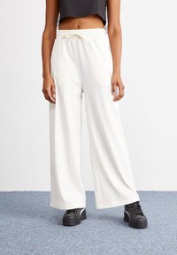 Žena v širokých bílých ribbových kalhotách s páskem na stažení a černých platformových teniskách stojící na koberci.