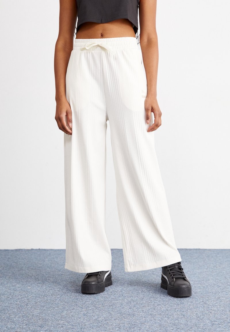 Žena v širokých bílých ribbových kalhotách s páskem na stažení a černých platformových teniskách stojící na koberci.