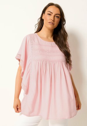 Femme portant un chemisier ample rose clair avec des détails en dentelle et des plis, associé à un pantalon blanc, debout devant un fond neutre.