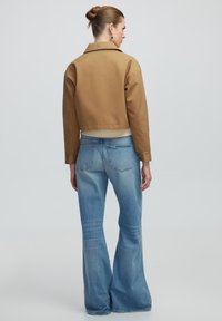 Kurtzbraune Jacke mit weit ausgeschnittenem Kragen, kombiniert mit hellblauen Schlagjeans, die über Gesäßtaschen und Auswaschungen verfügen.