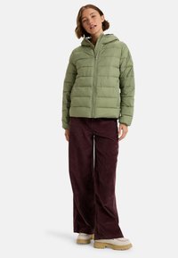 Chaqueta acolchada verde claro con capucha, con cremallera frontal y textura acolchada. Combinada con pantalones de pana burdeos oscuros y zapatos beis.