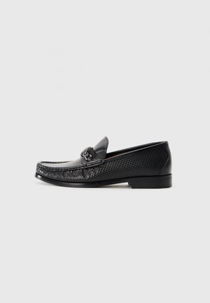 Black leather loafers with a textured woven design, featuring a metallic accent on the front, and a flat sole for ease of wear.