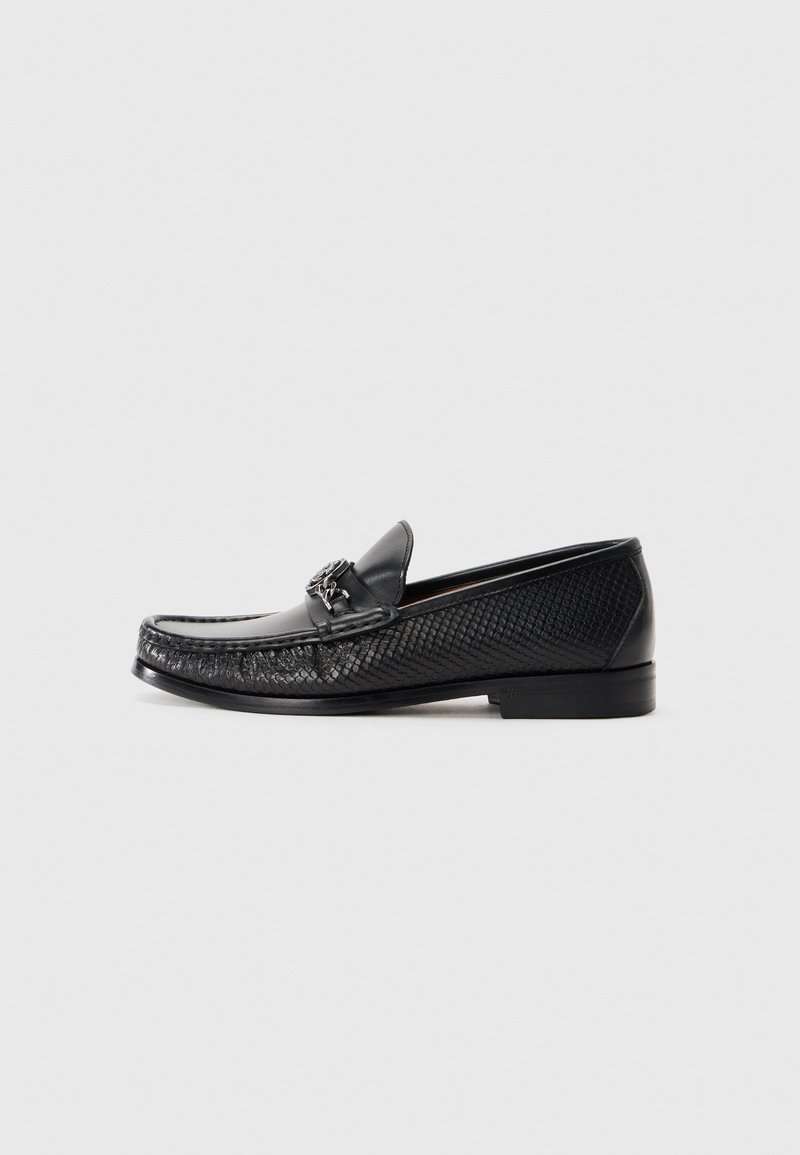 Schwarze Lederloafers mit einem strukturierten geflochtenen Design, ausgestattet mit einem metallischen Akzent auf der Vorderseite und einer flachen Sohle für ein angenehmes Tragegefühl.