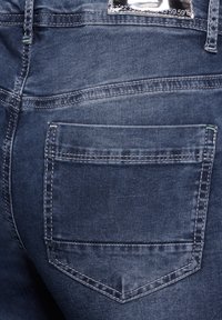 Pantalones vaqueros de mezclilla azul oscuro con costuras claras, que presentan un bolsillo trasero único con una solapa puntiaguda y una etiqueta encima.