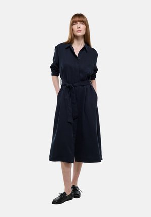 Femme aux cheveux longs et raides portant une robe midi noire à manches longues, ceinturée, avec des poches, et des chaussures noires à lacets, se tenant sur un fond blanc.