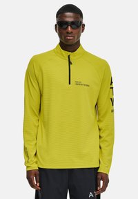 Jasnożółty sweter z długim rękawem i półzamkiem przy kołnierzu, teksturowany materiał, czarne akcenty oraz nadruki na klatce piersiowej i rękawie.