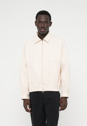 Lys beige lynlåsjakke med spredt krave, to for-lemme, elastisk talje og kontrasterende syninger. Blød stoftekstur.