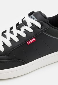 Baskets en cuir noir avec des lacets blancs et une semelle extérieure blanche. Elles présentent une petite étiquette rouge Levi's et de petites perforations sur les côtés.