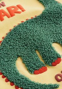 Grünes, strukturiertes Dinosaurierdesign mit flauschigem, geschlungenem Material auf einem gelben Hintergrund. Verfügt über rote Akzente und verspielten Text darüber.