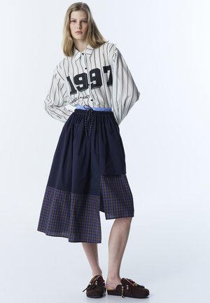 Mladá žena v pruhovaném tričku s nápisem „1997“, tmavě modré sukní s kostkovanými záplatami a hnědých nazouvacích botách s korálkovými střapci, stojící na bílém pozadí.