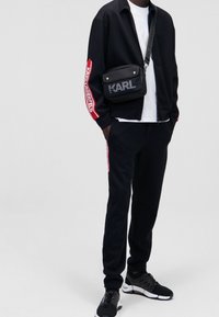 Chaqueta negra con detalles rojos y logo en las mangas, combinada con pantalones negros. Los accesorios incluyen un bolso cruzado negro con detalles de malla.