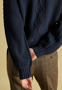 Jūras zila adīta džemperis ar teksturētu šuvi un virkņu rakstu priekšpusē, apvienots ar brūnām rūtainām biksēm.