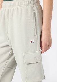Helle graue Jogginghosen aus weichem Material mit einem elastischen Bund, seitlichen Taschen und einer Cargo-Tasche mit genähtem Detail.