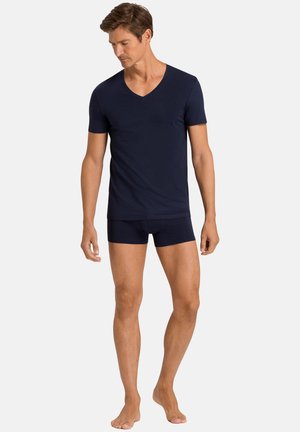 Uomo che indossa una maglietta scollo a V a maniche corte blu scuro e boxer coordinati, in piedi a piedi nudi davanti a uno sfondo semplice.