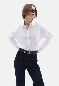 Witte overhemd met lange mouwen, gecombineerd met donkerblauwe high-waisted jeans en eenbruine riem. Accessoires zijn zwarte zonnebrillen.