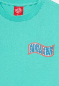 Turkis bomull T-skjorte med en rød etikett og et bølgete grafisk design med "SANTA CRUZ" i fete blå bokstaver. Glatt tekstur.