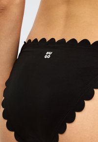 Black bikini bottom featuring a scalloped edge design, made from smooth fabric, with "HUGO" logo printed in white.