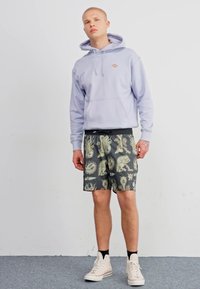 Sudadera de color púrpura claro con cordones, pantalones cortos grises con patrones que presentan diseños abstractos y zapatillas beige sobre un suelo de moqueta gris.