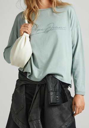 Femme portant une chemise à manches longues vert clair avec le texte "Pepe Jeans", tenant un sac blanc et une veste noire attachée autour de la taille.
