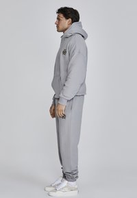 Felpa grigia con cappuccio e pantaloni abbinati realizzati in morbido tessuto, con un logo sul petto e tasche laterali sui pantaloni. Sneakers bianche.