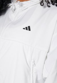 Personne portant une veste zippée blanche avec un logo triangulaire noir et des cordons près du cou, cheveux bouclés visibles sur le bord.