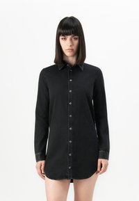Woman with straight dark hair wearing a long black buttoned shirt dress standing against a plain white background.