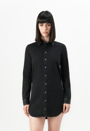 Woman with straight dark hair wearing a long black buttoned shirt dress standing against a plain white background.