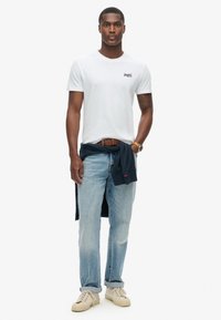 Hvid t-shirt med rund hals og lille logo, lyseblå denimjeans, marineblå sweatshirt bundet om livet og beige sneakers. Enkel, afslappet påklædning.