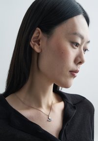 Sudraba kaklarota ar trīsstūra formas piekariņu, kam ir apļveida izgriezums, nēsāta uz melnas krekliņa ar apkakli. Gluda tekstūra, minimālisma dizains.