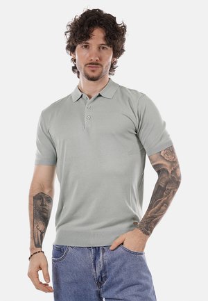 Uomo con capelli ricci che indossa una polo grigio chiaro e jeans blu, che mostra tatuaggi dettagliati su entrambi gli avambracci su uno sfondo semplice.