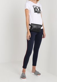 Svart bälteväska i konstläder bärs i midjan, vit t-shirt med svart tryck, marinblå leggings med röda sidostråk, och randiga sandaler.