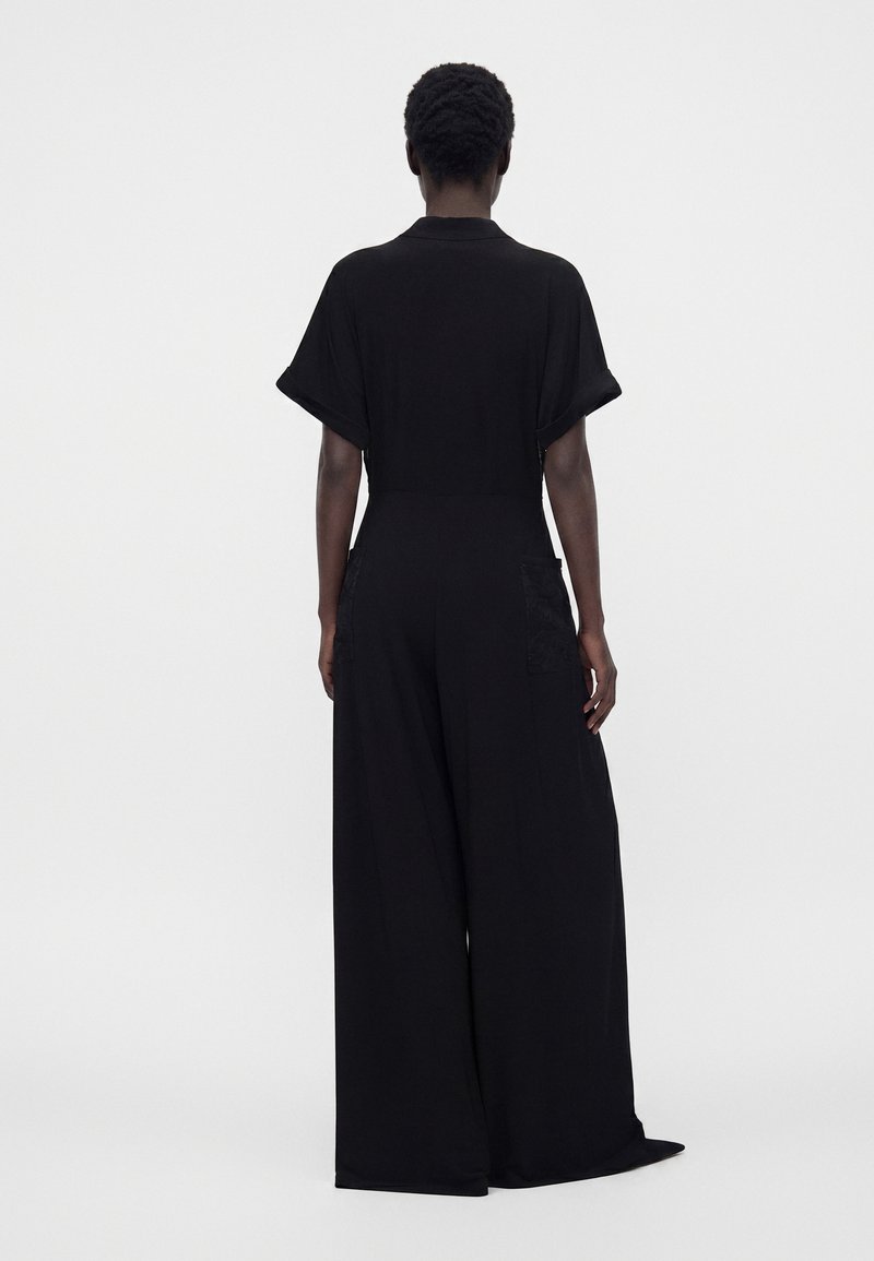 Personne vue de dos portant une combinaison noire à manches courtes avec jambes larges et deux poches arrière sur un fond clair uni.