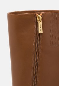 Botte en cuir marron à texture lisse, avec un détail de fermeture éclair dorée et des coutures verticales le long du côté. La tirette de la fermeture éclair porte le nom de la marque.