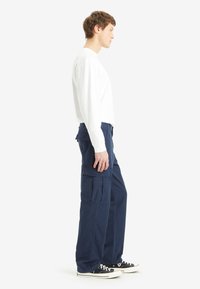 Navy cargo pants with multiple pockets, made of cotton. Paired with a white long-sleeve shirt and black sneakers.