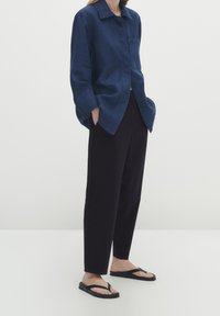 Marineblauwe linnen overhemd met een kraag en een oprolzakje aan de voorkant, gecombineerd met zwarte tapered broeken en zwarte teenslippers op een effen achtergrond.