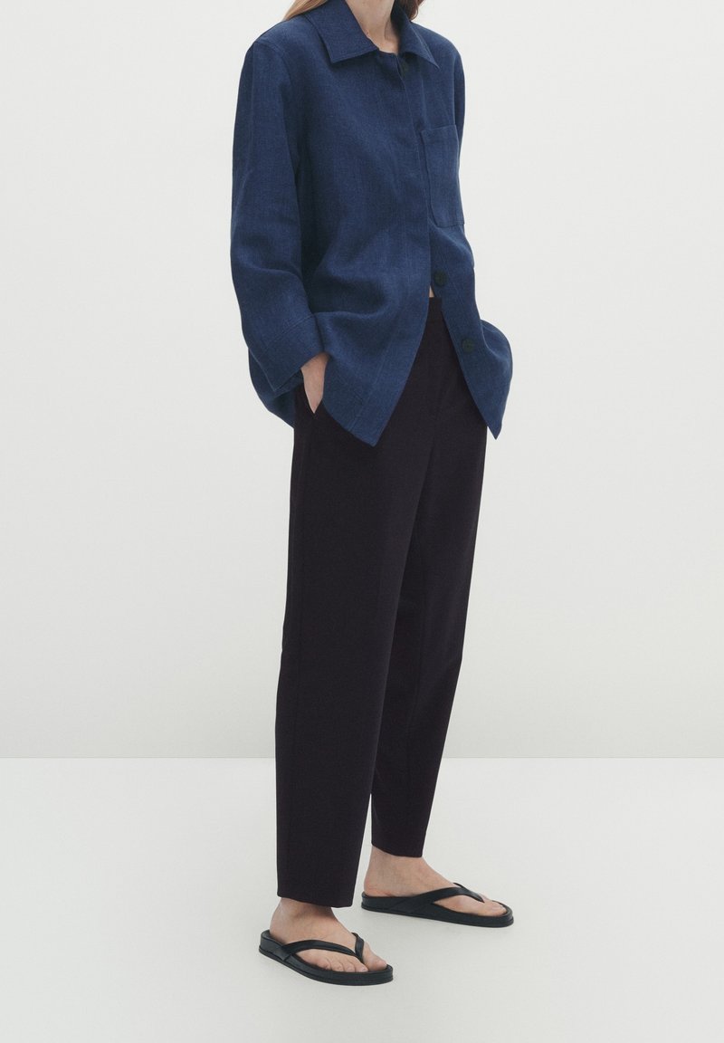 Marineblauwe linnen overhemd met een kraag en een oprolzakje aan de voorkant, gecombineerd met zwarte tapered broeken en zwarte teenslippers op een effen achtergrond.