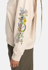 Felpa beige con illustrazioni floreali colorate e fantasiose sulla manica sinistra. Dotata di tasca a canguro e polsi a coste.