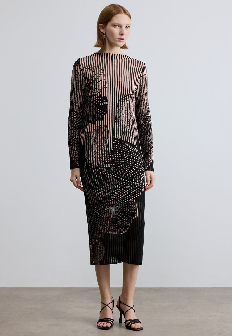 Robe mi-longue à rayures noires et nude avec motifs floraux, manches longues et silhouette droite. Associée à des talons noirs à brides.