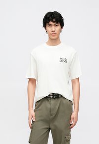 Hvid bomuld T-shirt med korte ærmer, med en sort "HAPPY HOLIDAYS" grafik på venstre bryst. Båret med grønne cargobukser og et sort bælte.