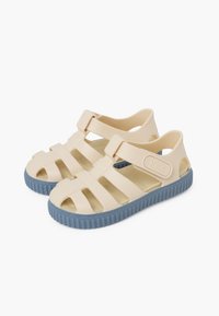 Beige rubber sandals with cut-out straps, adjustable buckle, and textured blue sole, featuring a logo on the side.