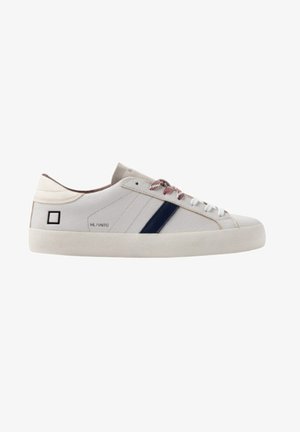 Witte lage sneaker met een marineblauwe zijstreep, rood-witte veters, een beige zool en een klein zwart vierkantje vlak bij de hiel.