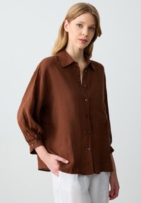 Camicia in lino marrone con colletto, chiusura a bottoni, taschino sul petto e maniche ampie. Indossata con pantaloni bianchi.
