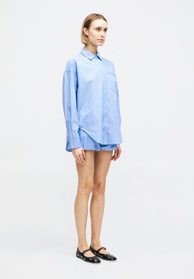 Νεαρή γυναίκα που στέκεται, φοράει υπερμεγέθη ανοιχτομπλε ριγέ πουκάμισο με κουμπιά, ταιριαστά σορτς και μαύρα παπούτσια Mary Jane με τρύπες για κορδόνια.
