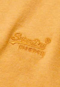 Gul bomuldsstof med en let struktureret overflade; har broderet "SuperDry" logo i matchende gul tråd.