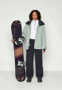 Snowboarder holding a dark snowboard with white bindings, wearing a light gray jacket, white top, and black pants in a neutral setting.
