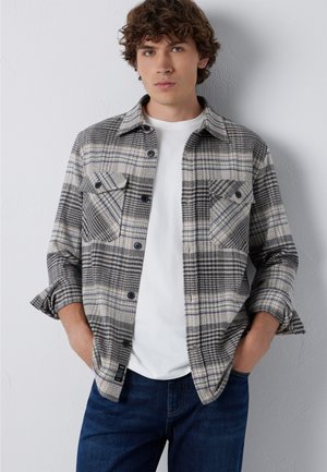 Junger Mann mit lockigem Haar, der ein grau-schwarz kariertes Hemd über einem weißen T-Shirt und blauen Jeans trägt, Hände in den Taschen.