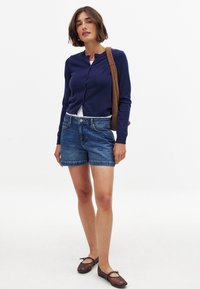 Mornarsko modri cardigan z okroglim izrezom in gumbi, v kombinaciji z svetlo modrimi jeans kratkimi hlačami. Rjava torba čez ramo in rjave balerinke.
