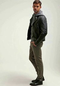 Veste en cuir noir de motard avec des détails matelassés, sweat à capuche gris en dessous, pantalon slim olive et bottines noires.