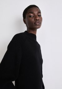 Pulôver preto de malha canelada com gola alta e mangas longas, apresentando costuras estruturadas nos ombros para um detalhe adicional.